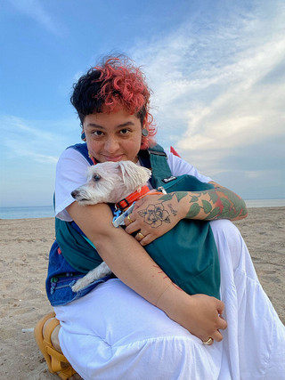 A photograph of a young person on a beach that kneels smiling in the sand while holding a small white dog with an orange collar. The person has gauged earrings, tattoos, orange hair, and wears a a white skirt, a denim vest, and orange sandals.